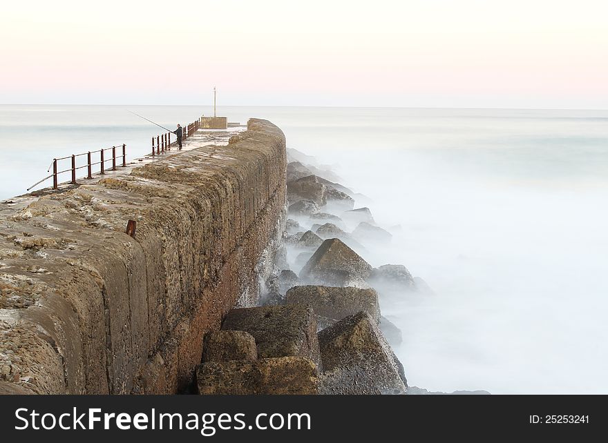 One solitary man is standing on a peer fishing just after sunset in Port Alfred, South Africa. It is a long exposure. One solitary man is standing on a peer fishing just after sunset in Port Alfred, South Africa. It is a long exposure.