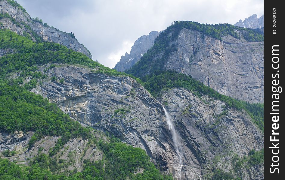 Alpine rocky walls with waterfall and surrounded by green trees. Alpine rocky walls with waterfall and surrounded by green trees.