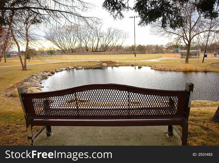 A picture of a park and pond with an empty bench. A picture of a park and pond with an empty bench