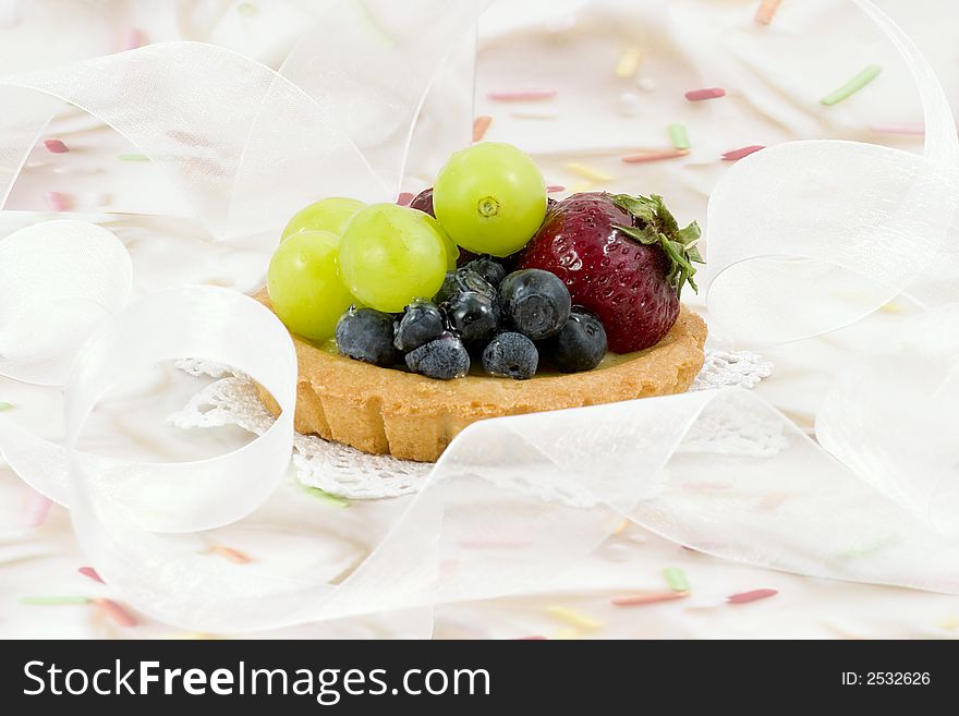 Fruit tart covered with strawberries,blueberries and green grapes on a doily in the middle of delicate white ribbon. The background is cake icing  with colored sprinkles. Fruit tart covered with strawberries,blueberries and green grapes on a doily in the middle of delicate white ribbon. The background is cake icing  with colored sprinkles.