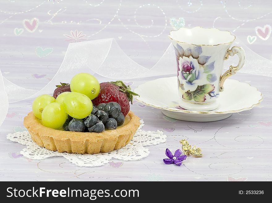 Fruit tart covered with strawberries,blueberries and green grapes on a doily with ribbon and a vintage tea cup. Fruit tart covered with strawberries,blueberries and green grapes on a doily with ribbon and a vintage tea cup.