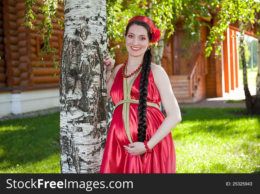 A happy young pregnant girl is standing under a tree in a red dress