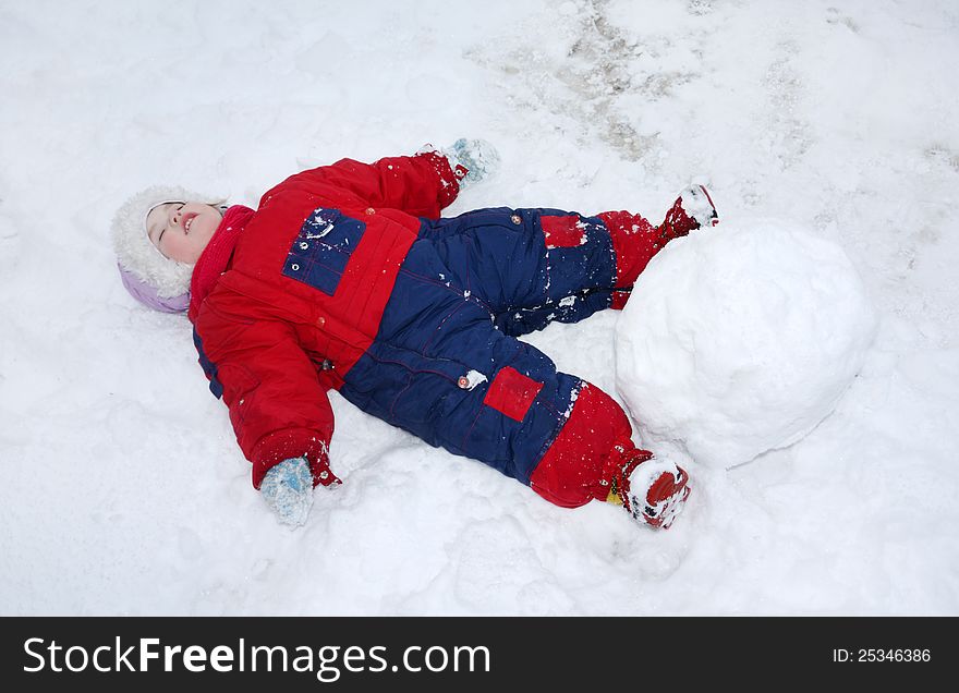 Little tired girl wearing warm jumpsuit lies on snow near big snowball. Little tired girl wearing warm jumpsuit lies on snow near big snowball