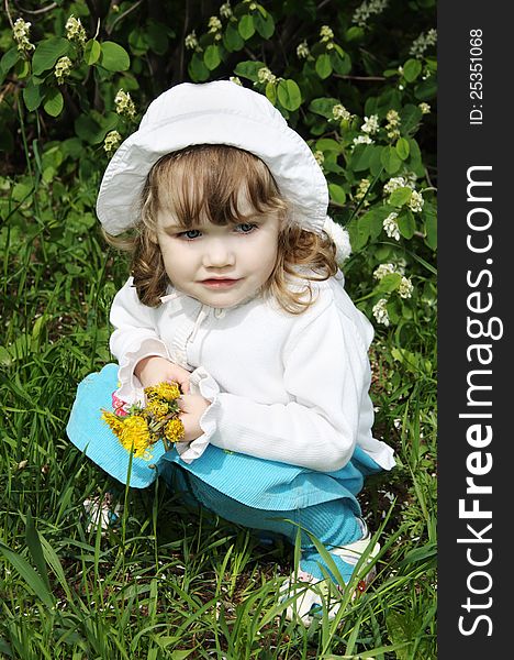 Beautiful little girl wearing white panama with yellow dandelions sits on grass and looks into distance. Beautiful little girl wearing white panama with yellow dandelions sits on grass and looks into distance