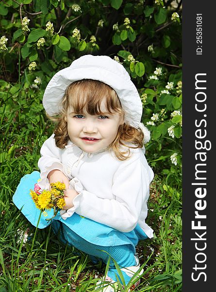 Beautiful little girl wearing white panama holds yellow dandelions and sits on grass. Beautiful little girl wearing white panama holds yellow dandelions and sits on grass