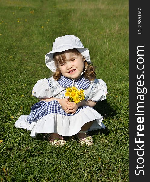 Beautiful little girl wearing dress with yellow dandelions sits on grass and blinks sun. Beautiful little girl wearing dress with yellow dandelions sits on grass and blinks sun