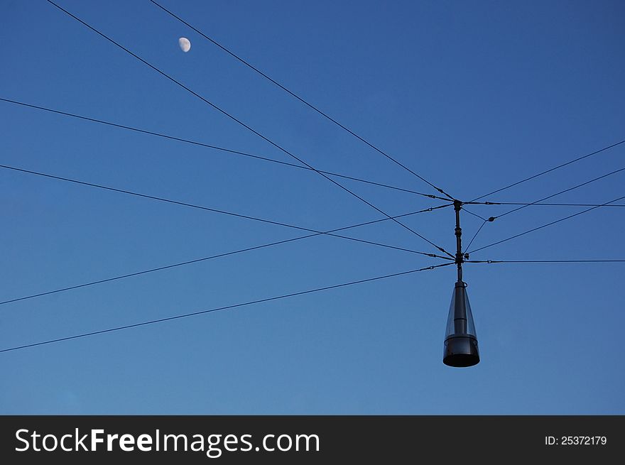 A free-hanging street lamp is seen against a deep blue evening sky with a small moon. A free-hanging street lamp is seen against a deep blue evening sky with a small moon