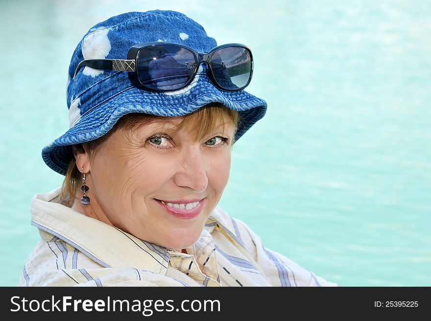 Adult woman in jeans panama and sunglasses resting near the water and looks at the camera smiling