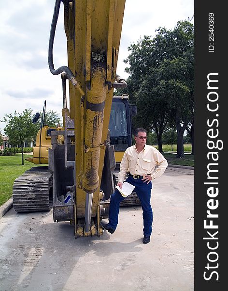 A man standing with his foot on the bucket of an idle backhoe with documents in his hand. A man standing with his foot on the bucket of an idle backhoe with documents in his hand.