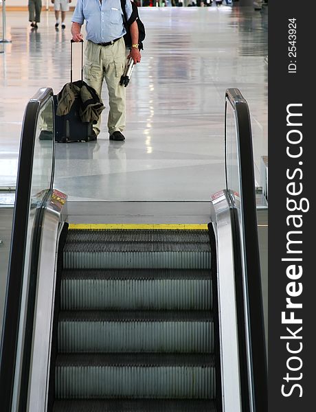 A bodyshot of a man standing by escalators in an airport with hand luggage. A bodyshot of a man standing by escalators in an airport with hand luggage