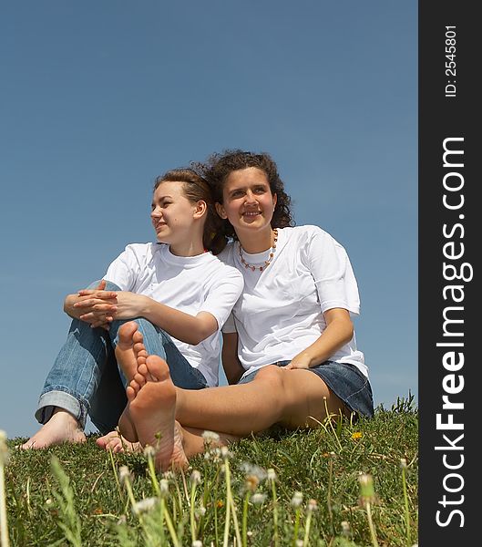 Two nice young girls sit on a grass on a background of the blue sky, smile and wave hands. Two nice young girls sit on a grass on a background of the blue sky, smile and wave hands