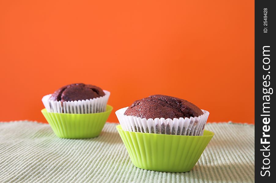 Two muffins in lime green plastic cups against orange wall on cotton table cloth. Focus on first muffin. Two muffins in lime green plastic cups against orange wall on cotton table cloth. Focus on first muffin