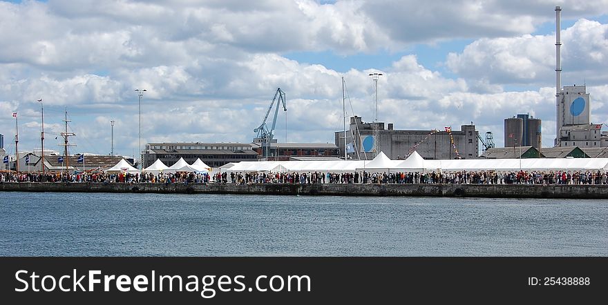 Crowds at a sailing festival in Aarhus, Denmark, gather along and in the white tents placed on the industrial harbor quay with silos and cranes. Crowds at a sailing festival in Aarhus, Denmark, gather along and in the white tents placed on the industrial harbor quay with silos and cranes.