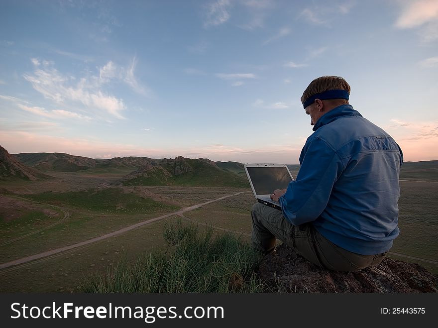 The man with the laptop sits on a hill against the evening sky