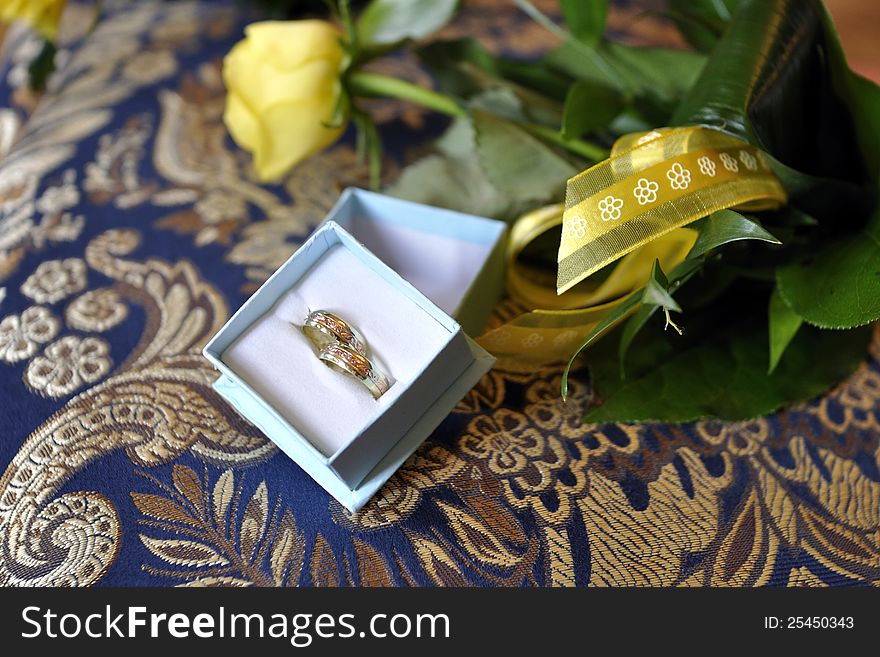 Pair of golden wedding rings in open box next to flowers on dark blue floral pillow background. Pair of golden wedding rings in open box next to flowers on dark blue floral pillow background