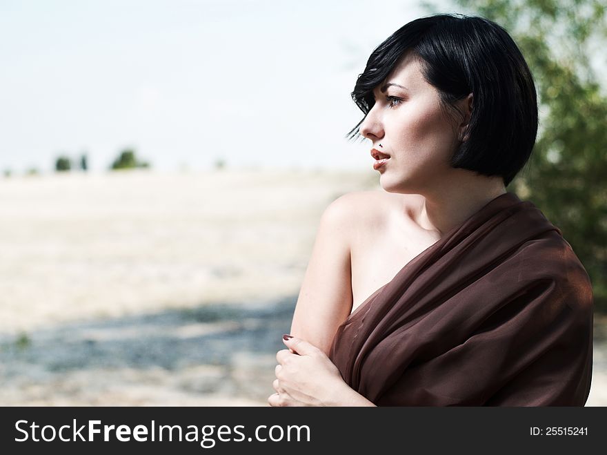 Beautiful black haired girl wrapped in brown cloth stands in the tree shade in a desert. Beautiful black haired girl wrapped in brown cloth stands in the tree shade in a desert