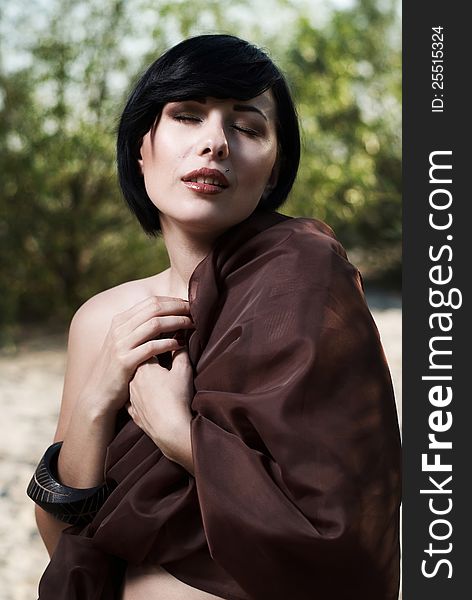 Beautiful black haired girl wrapped in brown cloth stands in the tree shade in a desert. Beautiful black haired girl wrapped in brown cloth stands in the tree shade in a desert