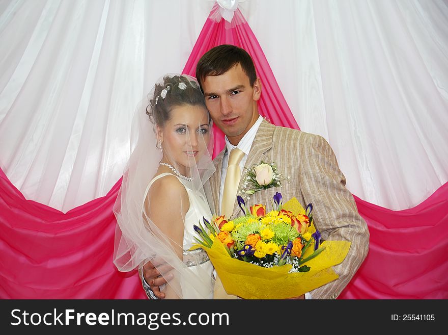 Happy groom with flowers hugs his beautiful bride near pink curtain indoor. Happy groom with flowers hugs his beautiful bride near pink curtain indoor