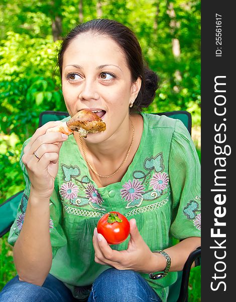 A young woman eating a chicken leg with a tomato. A young woman eating a chicken leg with a tomato