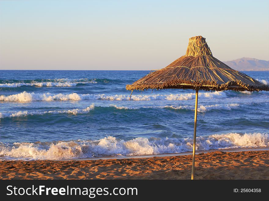 Umbrella from natural material on the cretan beach near the sea with waves on the sunset. Umbrella from natural material on the cretan beach near the sea with waves on the sunset