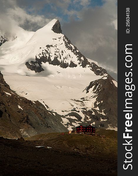 Fluhalp mountain hut under the massive mountain peak with cloudy sky.