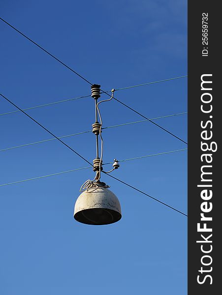 A free-hanging street lamp is seen against a blue sky with curled electrical wires connecting it to the supporting wires. A free-hanging street lamp is seen against a blue sky with curled electrical wires connecting it to the supporting wires