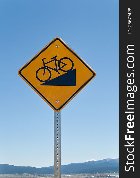 Steep hill biking warning sign against blue sky with mountains in the distant background. Steep hill biking warning sign against blue sky with mountains in the distant background