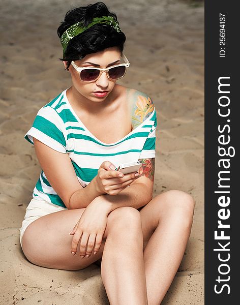 Pretty female model with tattoo and in sunglasses on the beach in the shade with phone in hands. Pretty female model with tattoo and in sunglasses on the beach in the shade with phone in hands