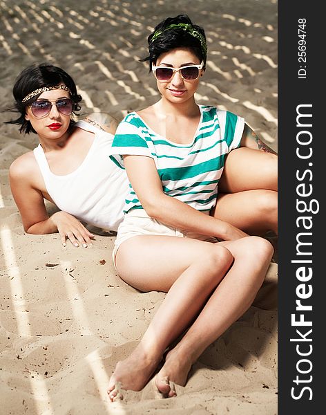 Two adorable women with tattoos wearing sunglasses on the beach in the shade