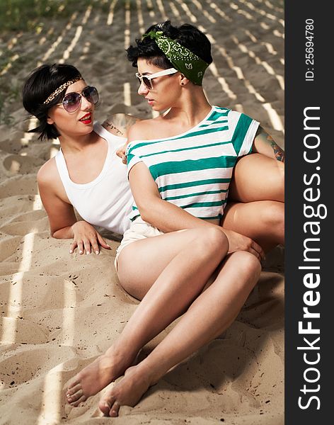 Two adorable women with tattoos wearing sunglasses on the beach in the shade