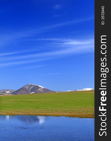 Early spring landscape with green field, lake, deep blue sky and snowy mountain