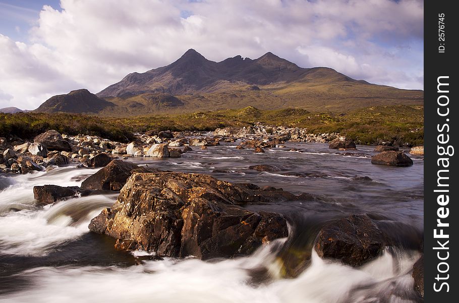 Morning sunlight lights the distant cullin hills in a warm glow. The photo is taken over a raging mountain stream with large rocks dominating the foreground. Morning sunlight lights the distant cullin hills in a warm glow. The photo is taken over a raging mountain stream with large rocks dominating the foreground