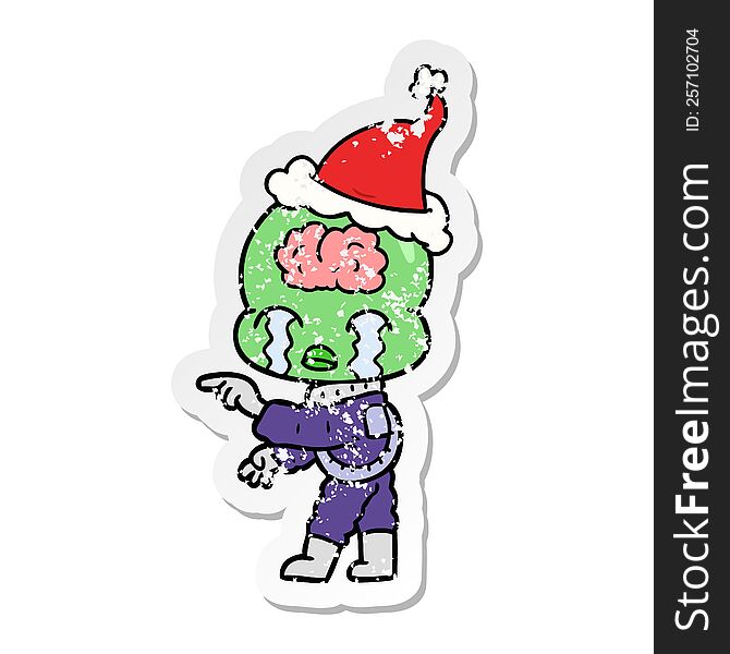 distressed sticker cartoon of a big brain alien crying and pointing wearing santa hat