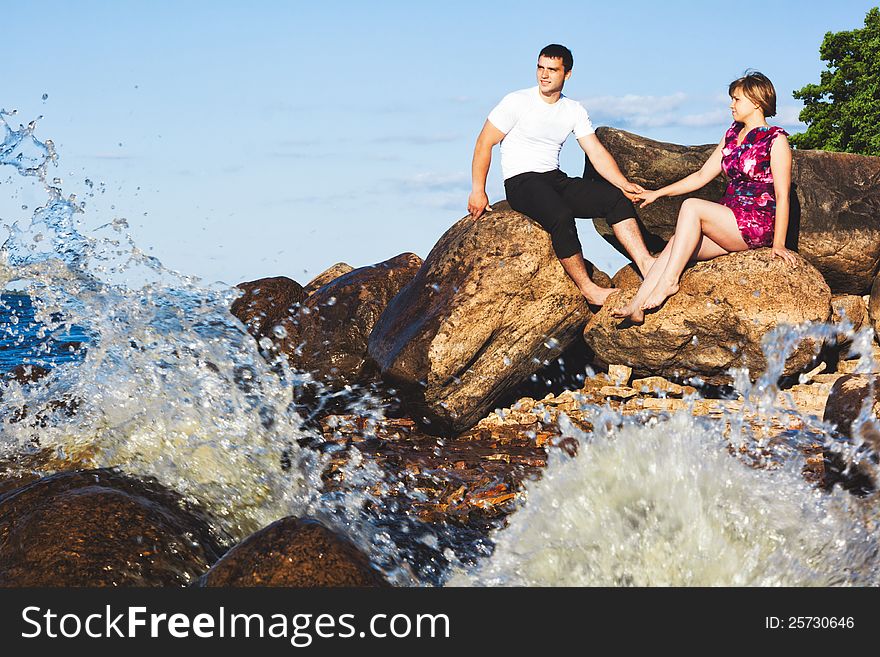 Couple holding hands, sitting on the shore of the lake, where the wave beats on the rocks. Couple holding hands, sitting on the shore of the lake, where the wave beats on the rocks