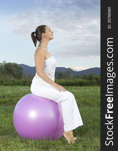 Cute young brunette girl performing yoga with a ball on green grass in park 
This photo  is part of a series of various yoga poses by this model.