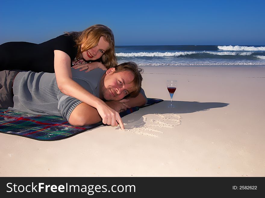 Young married couple enjoying their time on the beach. The lady is lying on her husband's back and drawing on the sand. Young married couple enjoying their time on the beach. The lady is lying on her husband's back and drawing on the sand.