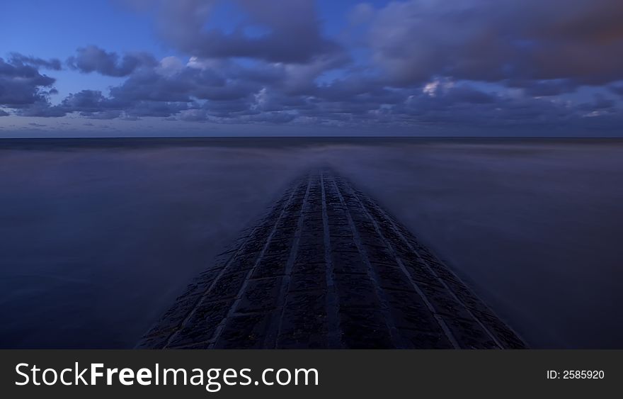 Stone breakwater at Belgian coast at a semi clouded night with a blue-isch atmosphere and milky water due to long exposure