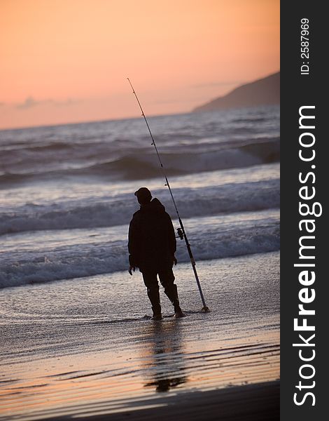Silhouette of man fishing in the ocean from the beach at sunset.