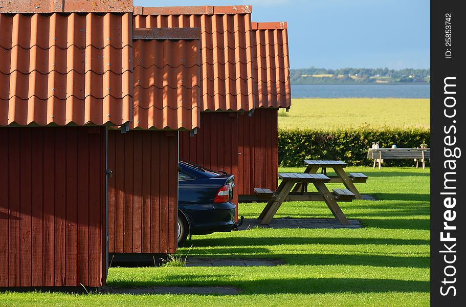 Four red holiday cottages with red tiled roofs form a line pointing to the bay behind a field. A car is parked between them and benches with tables are in front. The evening sun creates long shadows in the grass. Four red holiday cottages with red tiled roofs form a line pointing to the bay behind a field. A car is parked between them and benches with tables are in front. The evening sun creates long shadows in the grass.