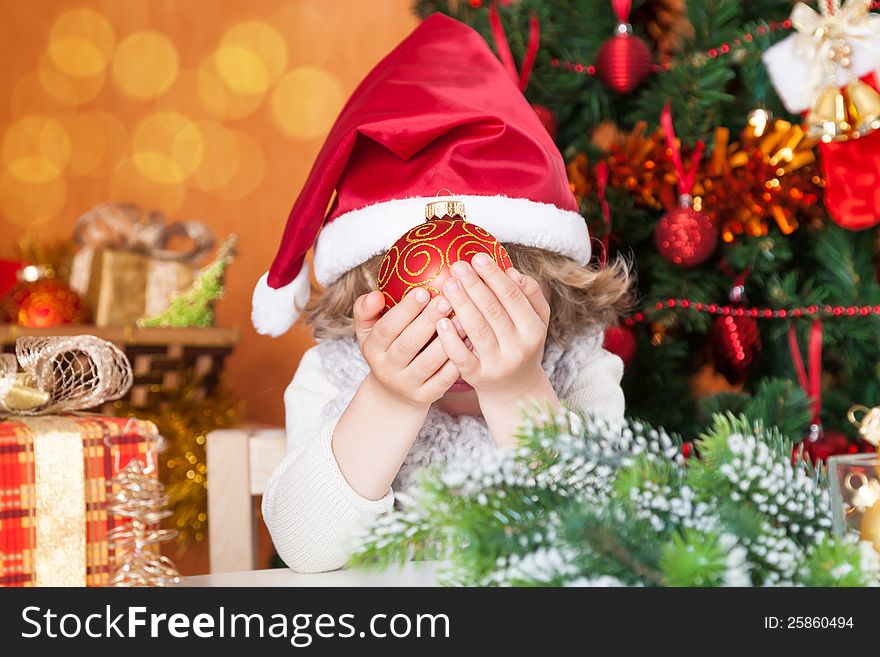 Happy baby in Santa`s hat hiding behind a ball against Christmas tree with decorations. Happy baby in Santa`s hat hiding behind a ball against Christmas tree with decorations