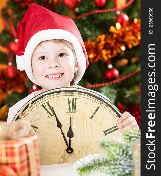 Happy kid in Santa`s hat holding old wooden clock against decorated Christmas background. Happy kid in Santa`s hat holding old wooden clock against decorated Christmas background