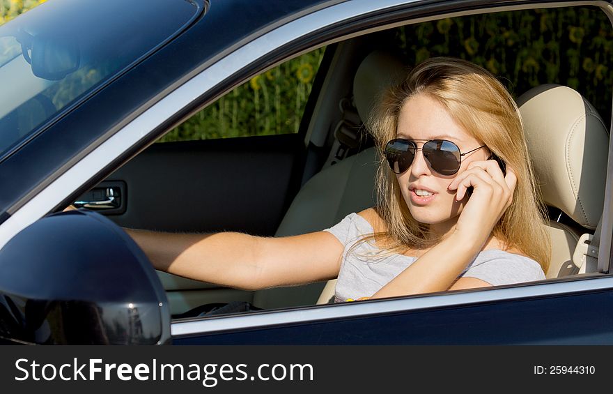 Young woman driver in sunglasses talking on her mobile phone while seated in the drivers seat of a stationary car. Young woman driver in sunglasses talking on her mobile phone while seated in the drivers seat of a stationary car