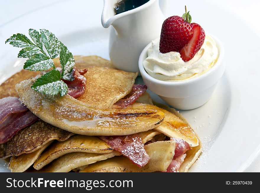 Stack of breakfast pancakes with bacon and banana. A jug of maple syrup and pot of cream accompanies it
