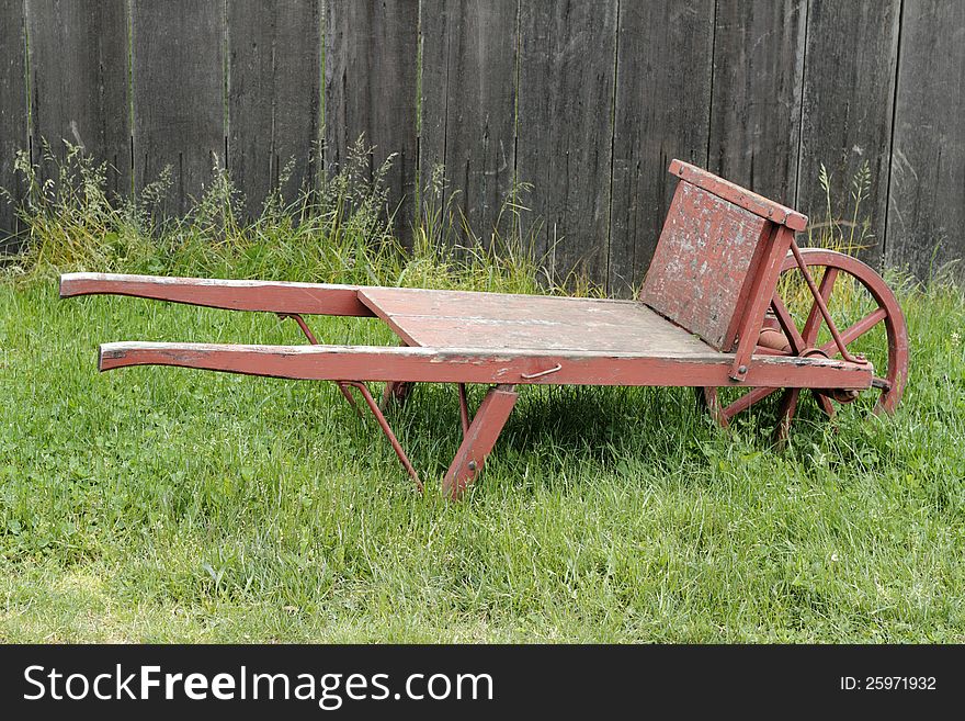 Very old wheel barrow made of wood and metal, painted red, sitting in a grassy field in front of an old wood fence. Very old wheel barrow made of wood and metal, painted red, sitting in a grassy field in front of an old wood fence.