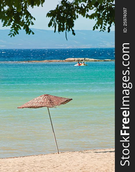 Vertical photo of a tropical holiday scene. Two chairs and a wooden mat umbrella is near the green / blue sea and beach. There are dark leaves on the upper side, creating a shady feeling. Vertical photo of a tropical holiday scene. Two chairs and a wooden mat umbrella is near the green / blue sea and beach. There are dark leaves on the upper side, creating a shady feeling.