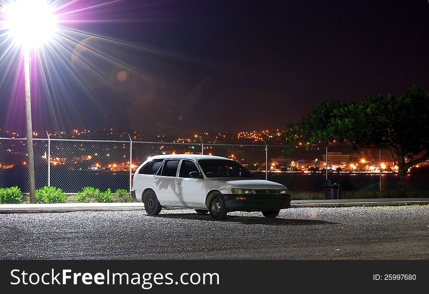 Cityscape at night with lights in the background and a car being bathed by a street light.