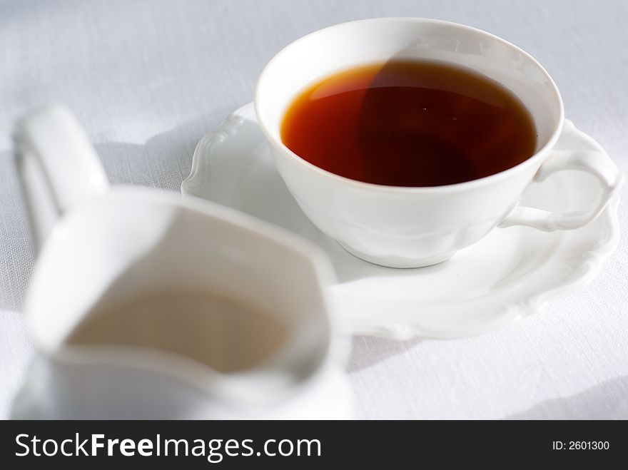 Cup of tea (focus on it) and porcelain milk jug on the white tablecloth. Shallow depth of field. Cup of tea (focus on it) and porcelain milk jug on the white tablecloth. Shallow depth of field.