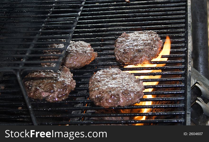 Hamburgers sit cooking with fire on a grill . Hamburgers sit cooking with fire on a grill .