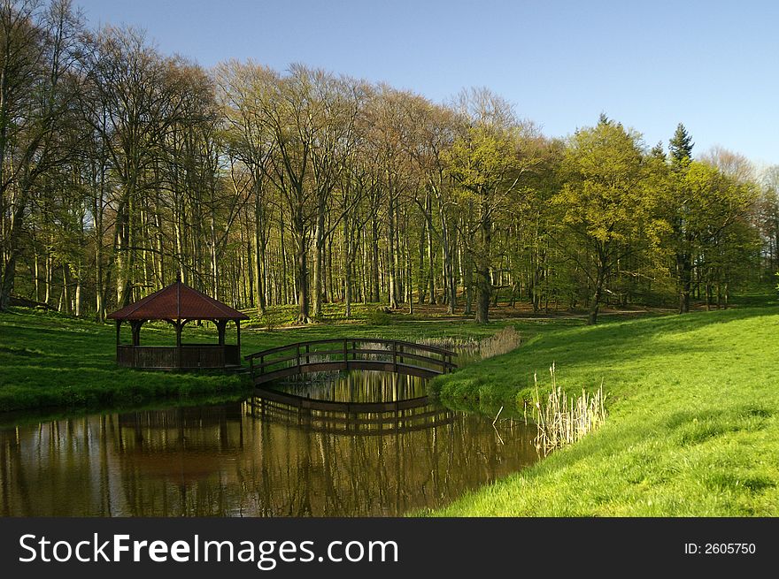 small wooden bridge and alcove in the park with fresh green trees, grass on the blue sky. small wooden bridge and alcove in the park with fresh green trees, grass on the blue sky