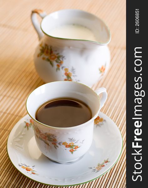 Cup of coffee or black tea (on the front, focus on the liquid surface) and a floral pattern porcelain milk jug on the wooden mat behind. Shallow DOF. Cup of coffee or black tea (on the front, focus on the liquid surface) and a floral pattern porcelain milk jug on the wooden mat behind. Shallow DOF.